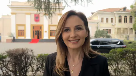 Josie Hannett / BBC Jorida Tabaku, seorang anggota parlemen dari Partai Demokrat, berdiri di luar Gedung Parlemen Albania di Tirana.