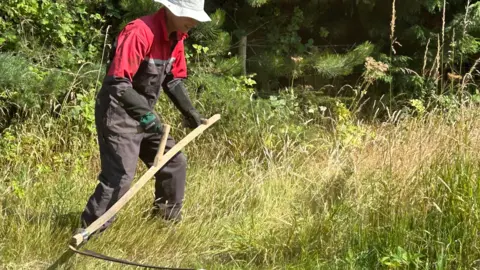 A man wearing a black and red jumpsuit, black protective gloves and a cream bucket hat, holding a large wooden and metal scythe, cutting long grass.