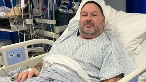 Damien Jeffries sits in a hospital bed wearing a hospital gown. He has cannulas in his hands and tattoos on his arms. He has short dark hair and stubble flecked with grey. 