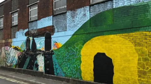 Chantelle Roberts colorful figures including patches of green and yellow pain on a dark brown brick building.