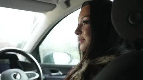 Alisa sitting in the driver's seat of a car. She has long dark hair and is wearing a furry coat