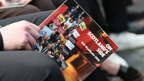 Close-up of a person holding a glossy booklet titled ‘Scotland on SNP Side – SNP Manifesto 2026,’ showing a collage of people standing on a stage, with the booklet resting on the person’s lap and other seated attendees blurred in the background