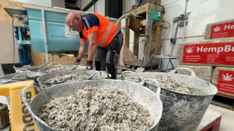 In a factory, wet hempcrete has been poured into several big buckets. It is a grey, fiberous mixture - like museli and yoghurt. In the background are bales of hemp shiv, wrapped in plastic with HempBuild written on them with a cannabis leaf logo.