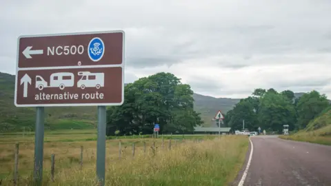 Getty Images A brown sign for the North Coast 500 route. There are direction pointers and illustrations of a car pulling a caravan, and a campervan. The sign also has the words: "NC500" and "alternative route". The sign is next to a rural road with trees and a hill in the distance.