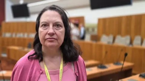 Paul Moseley/BBC A woman wearing a pink top and cardigan with a yellow lanyard stands in the foreground of a wood-panelled council or committee chamber. Curved desks, microphones, seating, and large display screens are visible in the softly blurred background.