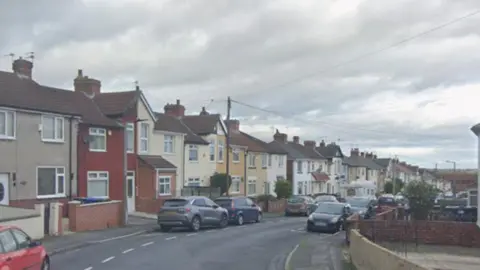A residential street lined with a long row of attached and semi-detached houses on both sides of the road. The homes are predominantly two-storey properties with pitched tiled roofs, brick chimneys, and small front gardens or paved areas enclosed by low brick walls and fences. In the foreground on the left, part of a red car is visible at the edge of the frame. Several other vehicles are parked along both sides of the street, including grey, blue, black, and silver cars. The road curves gently to the right as it continues into the distance. The houses vary slightly in appearance but share a similar style. Most have cream, white, beige, or light yellow façades, while one prominent house near the front is painted a dark red colour with white trim. Many of the homes have bay windows, small porches, and brick details around the entrances.