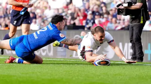 A man in a white England shirt dives over the line to score a try with a man in a blue shirt diving at him.