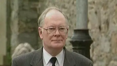 Lindsay Brown is a man with grey balding hair. He is wearing large round glasses and his lips are pursed. He is wearing a grey suit and a black tie.