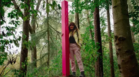  Elizabeth Blackie Artist Beccy McCray, with long brown hair, wearing a light-coloured jacket and trousers, stands next to a pink waymarker amid tall trees.