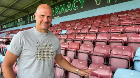 A bald man wearing a grey t-shirt with a pattern on the front is pictured pointing at claret and blue seats at Scunthorpe United's Glanford Park.