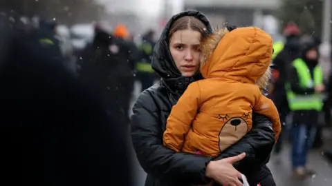 Reuters A woman in a black raincoat carrying a child at a border crossing near Ukraine. It is snowing. 