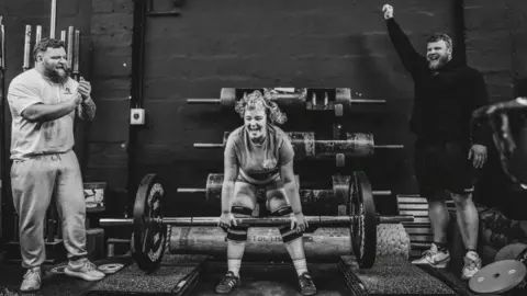 Gordon Siegal A black-and-white shot of lauri, laughing as she starts to lift a barbell with huge weights on each end. Her hair is in a curly ponytail and the Stoltman brothers, both strong-looking men with beards, are cheering her on from the sides.