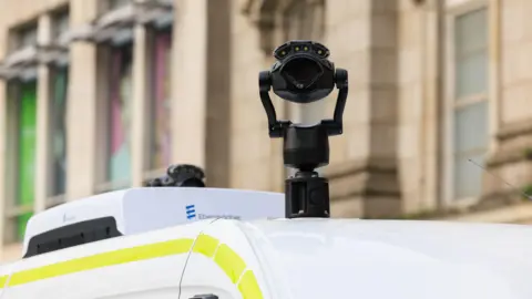 A close up of a black camera mounted on top of a police van. A blurred out building can be seen behind it.