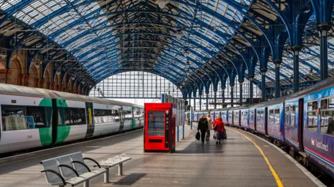A wide view of Brighton station, with a green and white Southern Train on one side of the platform and a blue and white Capital Connect train on the other side. A group of passengers are walking away towards the turnstiles, under the large blue steel and glass canopy.