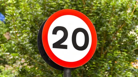 Stock image of a 20mph road sign. It is circular and has the number 20 in a white central section, surrounded by a red outline.