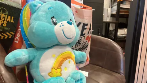 The image shows a light blue-coloured care bear with a shooting star on its stomach. It is also sat on a brown leather chair.