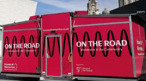 National Museums Liverpool A pink portacabin with the words On the Road - Immersive in the Community stands in a public area near on the Liverpool Pier Head