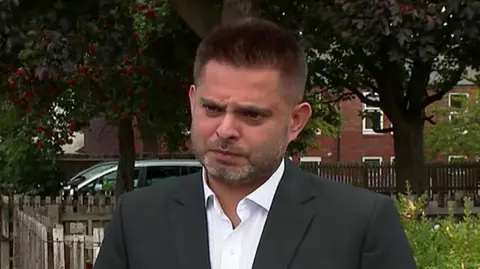 Waseem Zaffar a man in a black blazer and white shirt is looking at the camera with a stern expression