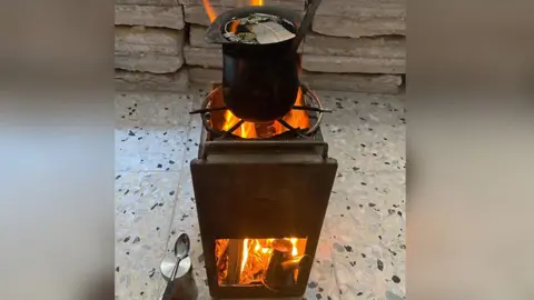 Asmaa Tayeh Rectangular black metal stove - with wood burning in bottom section and a pan on top. Placed on a tiled floor, with tongs and a spoon in front. 