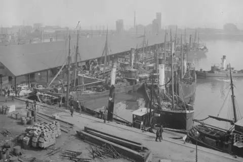 Topical Press/Getty Images A black-and-white photograph of numerous ships in a dock. About seven small trawlers with tall funnels are tied up in three rows at a quayside next to a long shed-like warehouse. A smaller vessel, possibly a tug, is at a right-angle to the trawlers. In the foreground, stacks of wood and other materials are on the quayside. In the background are the buildings of a town.