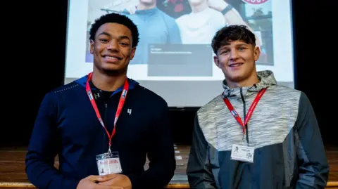 Two young men stand on a wooden stage in front of a large presentation screen. Both wear red lanyards with visitor badges. The screen behind them shows a photo of the same two men with another person outside a building.