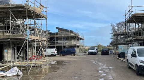 There is scaffolding around buildings on a construction site and several vans are parked nearby.