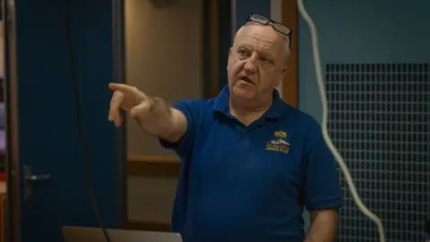 National Oceanography Centre Dr Bean Moat is wearing a blue t-shirt and pointing at something off camera with his right hand.