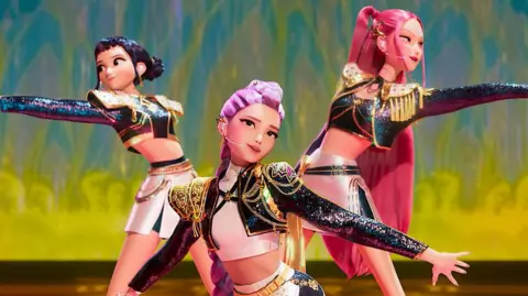 Netflix a aina no animācijas k -pop dēmonu medniekiem, kur trīs jaunas sievietes - Kpop elki - ir ģērbušās spīdīgos kostīmos un uzstājas uz skatuves. Viņi pozē ar izstieptām rokām.