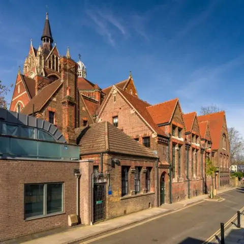 Historic England The side of a large red brick church with a tall ornate steeple and spire. The image is taken from the side of a narrow road. The sky is blue in the background. 