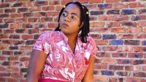 Family photograph Zanele Sibanda wearing a short-sleeved, pink patterned blouse and pink skirt. She is sitting in front of a brown brick wall and looking off to the left of the camera. Her black hair is twisted into shoulder-length locks with a few wooden beads secured at the top. 