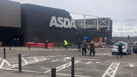 The outside of the Asda store in Hayle. A large section of the upper wall of the right-hand side of the modern-looking, two storey-high brown-bricked building is missing a large number of bricks, exposing a metal framework. The section is right beside the store's main "Asda" sign. Temporary fencing and red barriers have been put up around the damaged area. Several people are walking through the car park. A worker in a hi‑vis jacket is in the cordoned‑off section.