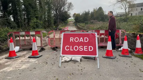 A wide shot of the bridge. At the front of the photo is a large red sign with white writing on it saying "Road closed". To each side of that is a row of traffic cones and behind it are three red and white plastic barriers, with large concrete blocks behind them. The damaged bridge can be seen in the distance. Dónal McAnallen stands to the right of the image in a maroon quarter-zip top and navy chinos. He has his left hand in his trouser pocket.