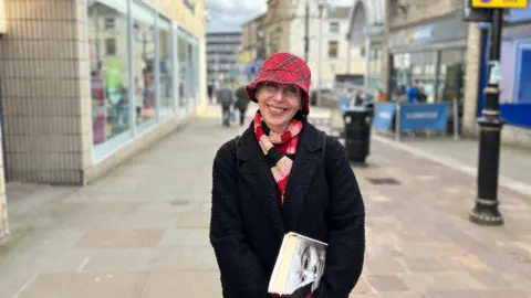 Amber Ward/BBC A woman in her seventies wearing a tartan hat and black coat. She is standing on a town high street and has a book under her arm. She is smiling at the camera.