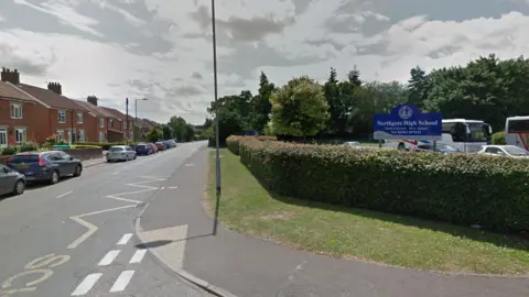 A road with cars parallel-parked along one side, to the left of the image, outside a row of houses, and to the right a hedge bordering a school car park, with a blue and white sign reading Northgate High School