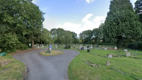 Google A screenshot from Google street view showing a cemetery with a road in the middle, with a small traffic island in the middle. There are trees on either edge of the cemetery and it's a cloudy day.