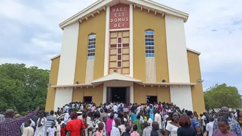 BBC/Nichola Mandil A congregration standing outside a church with the latin phrase Haec est domus Dei written on the front. The church is in the centre of the picture and the doors are open with people going through them. 