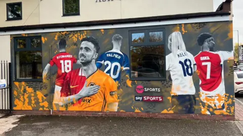 A mural of football players on a wall. It shows a football player with his hand on his chest, with his football shirt divided into two, representing two different clubs. Four other football players are behind him with their backs in view and not their faces.