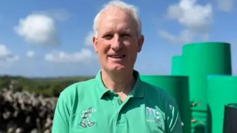Ray Cox wears a green polo shirt with the Parish Walk logo. He stands outside in sunshine in front of stone wall and has short white hair and is clean shaven. He is smiling for the camera.