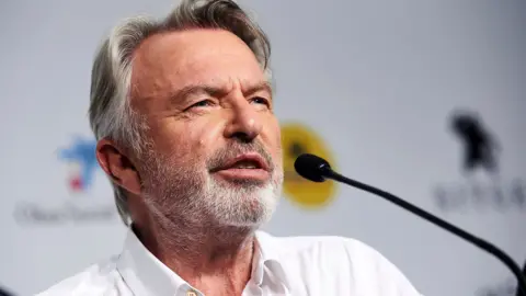 A head and shoulders crop of Neill looking away from camera while speaking into a microphone against a bokeh background, in Spain in 2019.