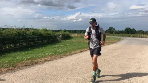 Katie Radley/BBC A man wearing a grey T-shirt, dark-coloured shorts, a dark grey cap with green writing, a dark grey running vest, trainers and green and orange long socks is running towards the camera. He is running on a road in the countryside with grass, hedges and trees behind him.