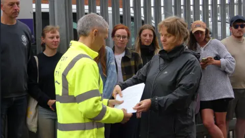 Port of Tyne Matt Beeton, an older man with short, grey hair, wearing a high-visibility coat. He is being handed the document by a woman with blonde-brown hair wearing a black, waterproof coat. Behind them, standing in front of a steel fence, is a row of campaigners in ordinary clothing.
