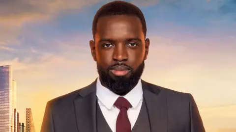 Naked A publicity image where Frederick Afrifa has a tidy dark beard and dark short hair. He is wearing a dark grey suit with a dark red tie and crisp white shirt. Behind him is a sunset or sunrise and part of the London skyline.