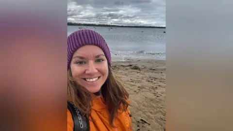 Mairead Morgan Mairead wears a cerise bobble hat and orange Dryrobe, and is standing on a beach. She is smiling.