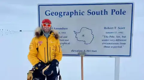 Jonny Huntington, in a yellow coat and sunglasses. He is wearing black trousers and black gloves. Behind him is a sign which says "Geographic South Pole" and behind that is snow. 