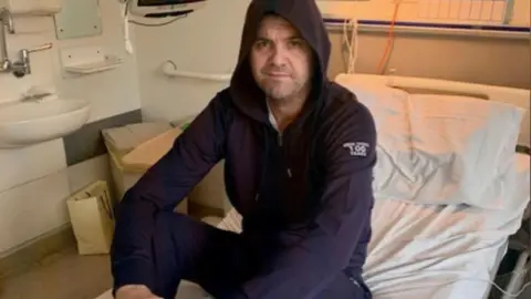 Family handout A man in a tracksuit sits on a hospital bed. 