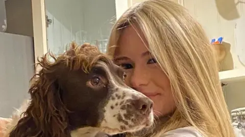 Tilly Bray. She has long, blonde hair and is holding a brown and white spaniel.