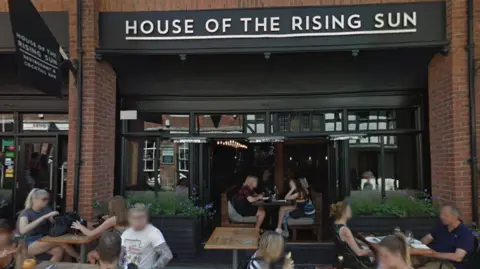 Google A bar with a black frontage and large bifold doors which are open, showing the inside where people are sitting at tables. Outside there are people sitting at wooden tables. A white sign reads "house of the rising sun"