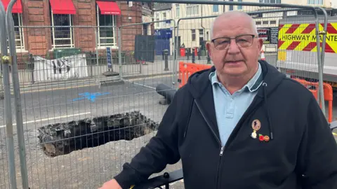 A grey-haired man wearing a jumper with military pins on it stands in front of a large rectangular-shaped sinkhole. The hole is surrounded by metal fencing. 