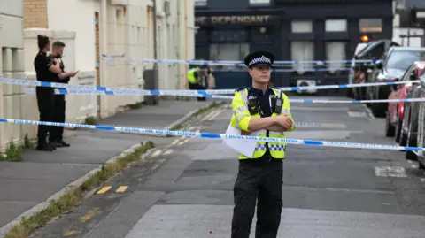Eddie Mitchell A street cordoned off by police. A police officer is stood directly behind the police tape cordon with his arms crossed. Another two officers are further into the cordon, stood on the left hand side.