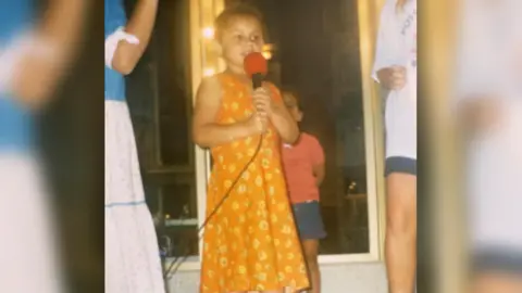 Aleighcia Scott Aleighcia singing when she was a child. She is wearing an orange dress and is holding a microphone. 